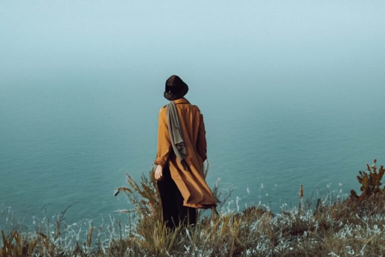 Person looking over to the sea, standing on a grassy cliff. The person has a hat on a jacket and las a large hbag over their shoulder