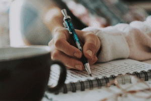 a hand holding a pen writing in a notebook. a coffee cup in the foreground